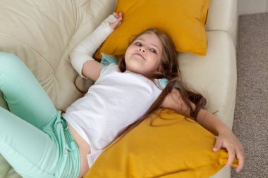 Kırık bileği ya da kolu olan bir çocuk gülümsüyor ve kanepede eğleniyor. Pozitif tutum, iyileşme ve çocuk konsepti.