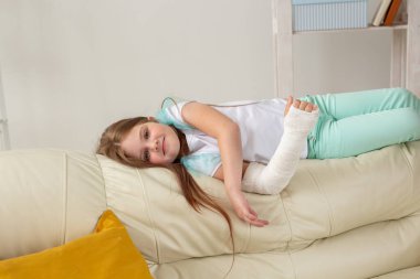 Kırık bileği ya da kolu olan bir çocuk gülümsüyor ve kanepede eğleniyor. Pozitif tutum, iyileşme ve çocuk konsepti.