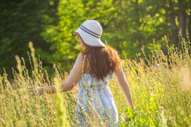 Güneşli yaz gününde kır çiçeklerinin arasında yürüyen güzel genç bir kadının arka manzarası. Yaz doğasıyla iletişim kurmanın zevki kavramı