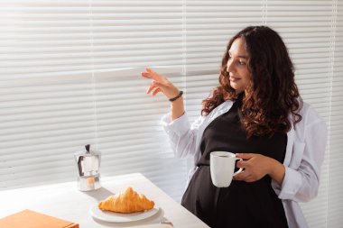 Neşeli, hamile, güzel bir kadın sabah kahvaltısı boyunca panjurlardan kahve ve kruvasanla bakıyor. Günaydın kavramı ve bir bebekle tanışmayı beklemek. Kopyalama alanı