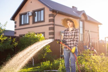 Beyaz kadın bahçıvan iş kıyafetleri içinde güneşli yaz gününde sebze bahçesindeki yatakları suluyor. Bahçede ve çiftlikte çalışma kavramı
