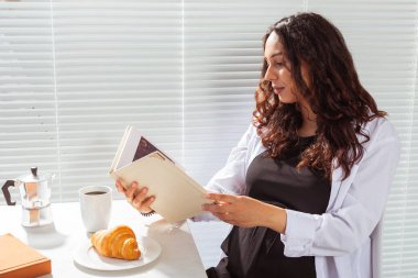 Genç ve mutlu bir kadının kitap okurken, arka planda kahve ve kruvasanla kahvaltı edişinin yan görüntüsü. Günaydın konsept ve keyifli öğle yemeği arası