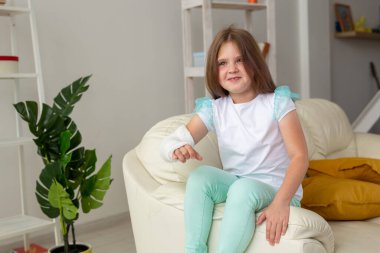 Kırık bileği ya da kolu olan bir çocuk gülümsüyor ve kanepede eğleniyor. Pozitif tutum, iyileşme ve çocuk konsepti.