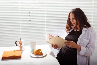 Genç ve mutlu bir kadının kitap okurken, arka planda kahve ve kruvasanla kahvaltı edişinin yan görüntüsü. Günaydın konsept ve keyifli öğle yemeği arası