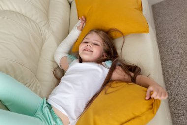 Kırık bileği ya da kolu olan bir çocuk gülümsüyor ve kanepede eğleniyor. Pozitif tutum, iyileşme ve çocuk konsepti.