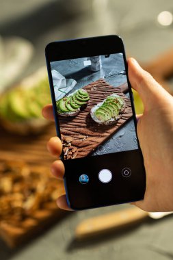 Eller, masada duran iki güzel sağlıklı ekşi krema ve avokado sandviçinin resimlerini akıllı telefondan çeker. Sosyal medya ve gıda kavramı