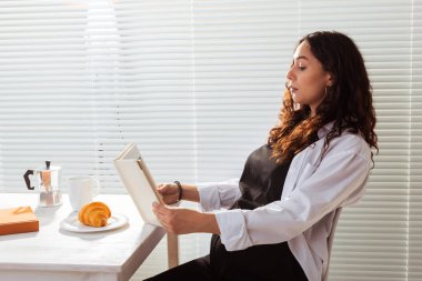 Genç ve mutlu bir kadının kitap okurken, arka planda kahve ve kruvasanla kahvaltı edişinin yan görüntüsü. Günaydın konsept ve keyifli öğle yemeği arası