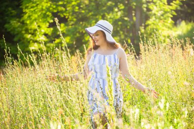 Uzun şapkalı ve elbiseli mutlu genç kadın güneşli bir günde yaz ormanında yürürken ellerini bitkilere doğru çekiyor. Yaz neşesi konsepti