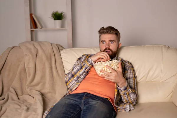 Bearded man watching film or sport games TV eating popcorn in house at night. Cinema, championship and leisure concept.