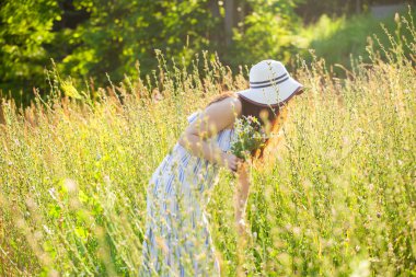 Uzun şapkalı ve elbiseli mutlu genç kadın güneşli bir günde yaz ormanında yürürken ellerini bitkilere doğru çekiyor. Yaz neşesi konsepti