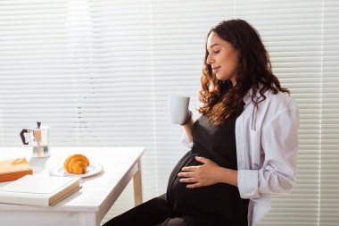 Genç ve mutlu bir kadının kitap okurken, arka planda kahve ve kruvasanla kahvaltı edişinin yan görüntüsü. Günaydın konsept ve keyifli öğle yemeği arası
