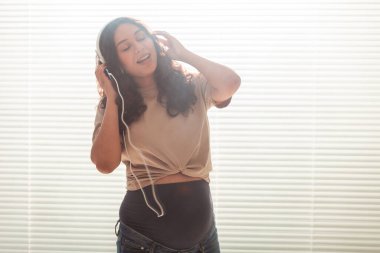 Kıvırcık saçlı esmer hamile kadın akıllı telefon ve kulaklık kullanarak hoş klasik müzik dinliyor. Bebekle tanışmadan önce rahatlatıcı ruh hali kavramı. Kopyalama alanı