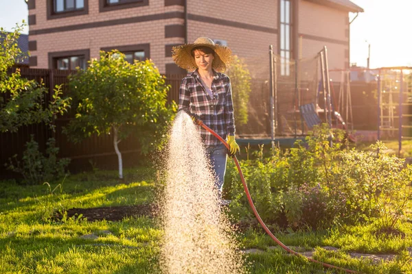 Donna felice giardiniere in abiti da lavoro innaffiando i letti nel suo orto nella soleggiata calda giornata estiva. Concetto di lavoro nell'orto della vostra azienda agricola — Foto Stock