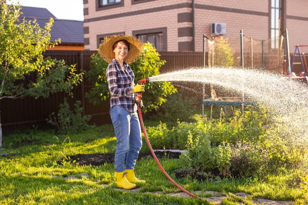 Donna felice giardiniere in abiti da lavoro innaffiando i letti nel suo orto nella soleggiata calda giornata estiva. Concetto di lavoro nell'orto della vostra azienda agricola — Foto Stock