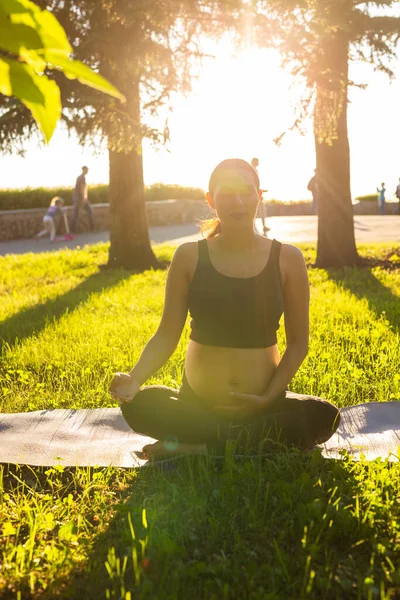 Lugn ung positiv gravid kvinna i gymnastik kostym gör yoga och meditera sitter på matta på grönt gräs på solig varm sommardag. Begreppet förberedelse för förlossning och positiv inställning — Stockfoto