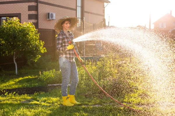 Donna felice giardiniere in abiti da lavoro innaffiando i letti nel suo orto nella soleggiata calda giornata estiva. Concetto di lavoro nell'orto della vostra azienda agricola — Foto Stock