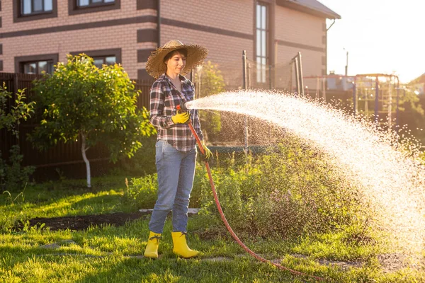 Donna giardiniere in abiti da lavoro innaffiare i letti nel suo orto nella soleggiata calda giornata estiva. Concetto di lavoro in giardino e nella tua azienda agricola — Foto Stock