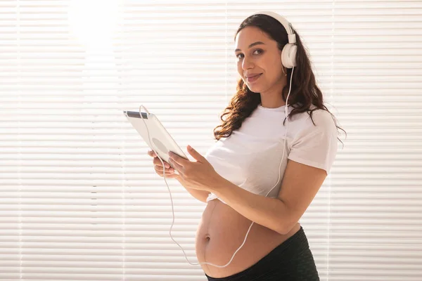 Красивая молодая беременная женщина выбирает музыку по подписке и слушает ее по утрам через наушники и планшеты. Концепция льгот для развития плода — стоковое фото