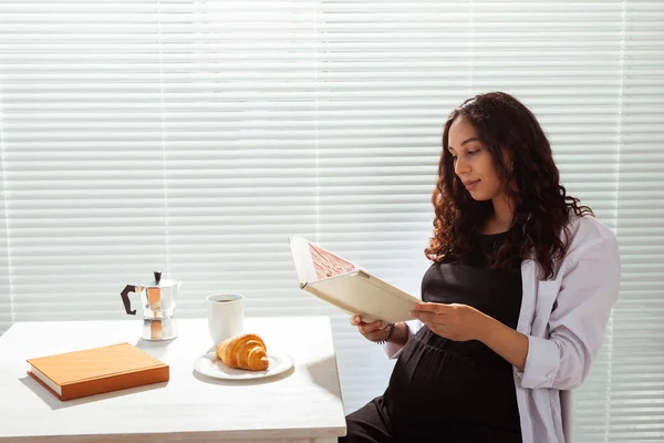 Genç ve mutlu bir kadının kitap okurken, arka planda kahve ve kruvasanla kahvaltı edişinin yan görüntüsü. Günaydın konsept ve keyifli öğle yemeği arası — Stok fotoğraf