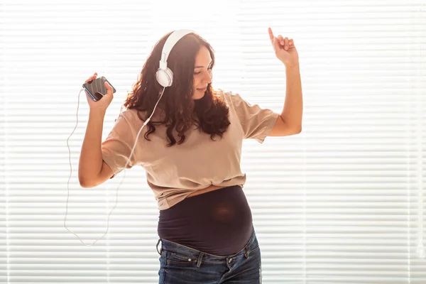 Curly-haired brunette pacified pregnant woman listens to pleasant classical music using smartphone and headphones. Concept of a soothing mood before meeting baby. — Stock Photo, Image
