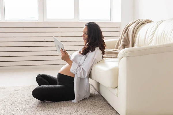 Gravid kvinna som håller tablett sitter på en matta nära en soffa i vardagsrummet hemma — Stockfoto
