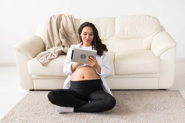 Gravid kvinna som håller tablett sitter på en matta nära en soffa i vardagsrummet hemma — Stockfoto