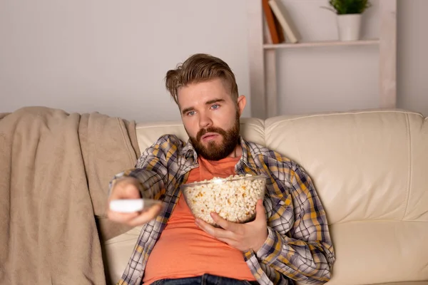 Πορτρέτο νεαρός άνδρας κάθεται στον καναπέ και βλέποντας τηλεόραση, κρατώντας τηλεχειριστήριο και τρώει ποπ κορν — Φωτογραφία Αρχείου