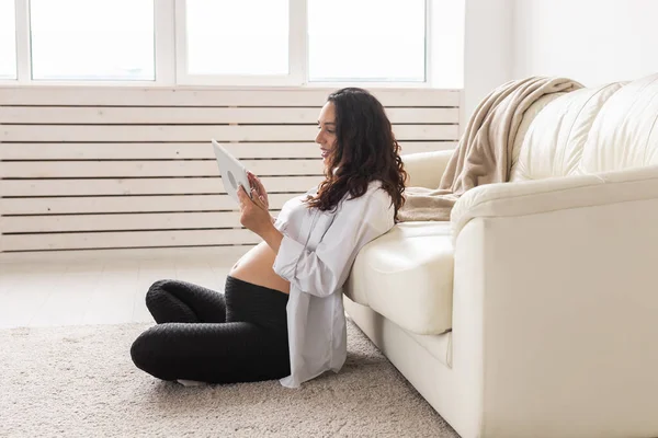 Femme enceinte tenant une tablette assise sur un tapis près d'un canapé dans le salon à la maison — Photo