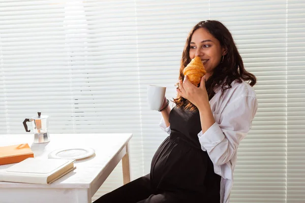 Felice incinta giovane bella donna mangiare croissant durante la colazione del mattino. Concetto di piacevole mattina e atteggiamento positivo durante la gravidanza — Foto Stock