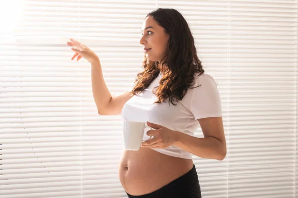 Curiosa jovem mulher grávida bonita bebendo chá e olhando através das persianas na janela. Conceito de alegria e boas notícias enquanto espera pelo bebê — Fotografia de Stock