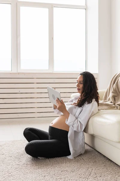 Gravid kvinna som håller tablett sitter på en matta nära en soffa i vardagsrummet hemma — Stockfoto