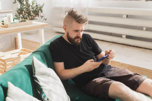 Χαλαρός άνθρωπος χρησιμοποιώντας ένα smartphone κάθεται σε πράσινο καναπέ στο σαλόνι στο σπίτι — Φωτογραφία Αρχείου