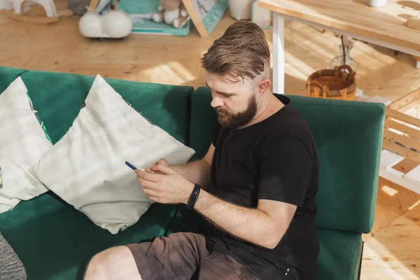 Hombre relajado usando un teléfono inteligente sentado en un sofá verde en la sala de estar en casa — Foto de Stock