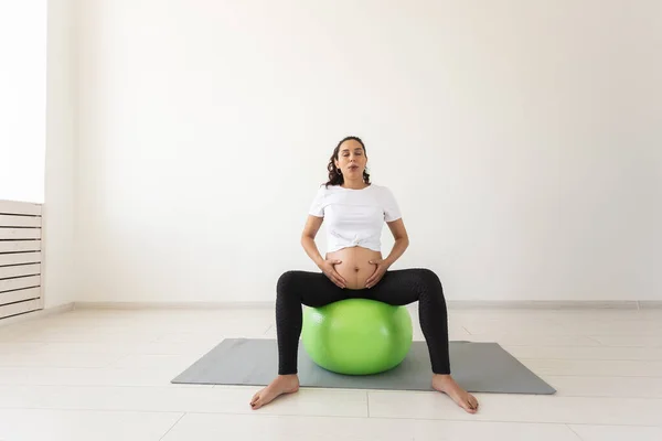 Una joven embarazada haciendo ejercicio de relajación usando una pelota de fitness mientras está sentada en una esterilla. Espacio de copia. —  Fotos de Stock