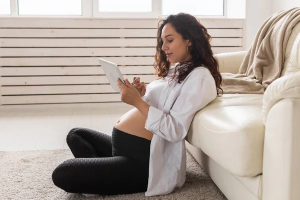 Gravid kvinna som håller digital medan tablett sitter på en matta nära en soffa i vardagsrummet hemma — Stockfoto