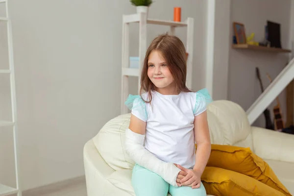 Niño con un yeso en una muñeca o brazo roto sonriendo y divirtiéndose en un sofá. Actitud positiva, recuperación y concepto de niño. — Foto de Stock