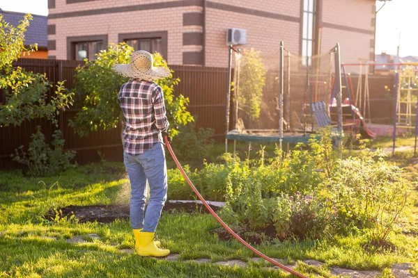 Donna felice giardiniere in abiti da lavoro innaffiando i letti nel suo orto nella soleggiata calda giornata estiva. Concetto di lavoro nell'orto della vostra azienda agricola — Foto Stock