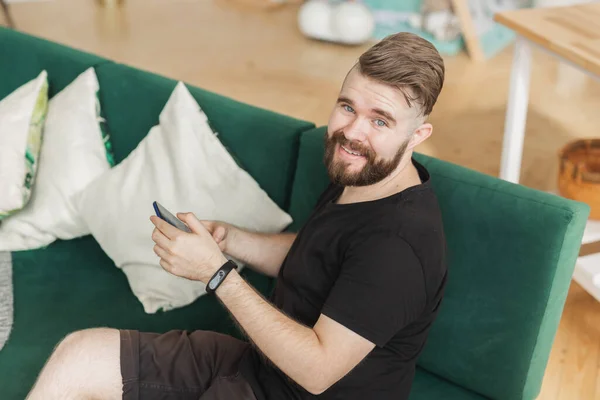 Hombre relajado usando un teléfono inteligente sentado en un sofá verde en la sala de estar en casa — Foto de Stock