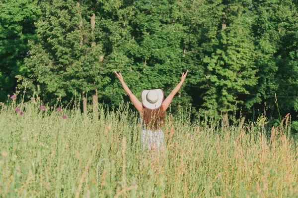 Vista trasera de la joven feliz con el pelo largo en el sombrero y el vestido tira de sus brazos hacia arriba mientras camina a través del bosque de verano en un día soleado. Concepto de alegría de vida — Foto de Stock