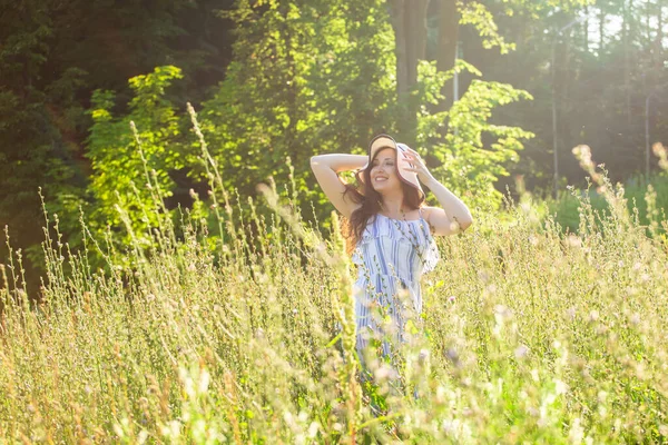 Giovane donna felice con i capelli lunghi in cappello e vestito a piedi attraverso la foresta estiva in una giornata di sole. Concetto gioia estiva — Foto Stock