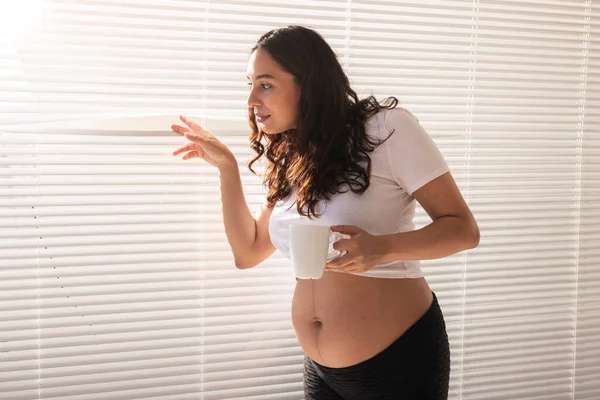 Любопытная молодая красивая беременная женщина пьет чай и смотрит сквозь жалюзи в окно. Радости и хороших новостей в ожидании ребенка — стоковое фото