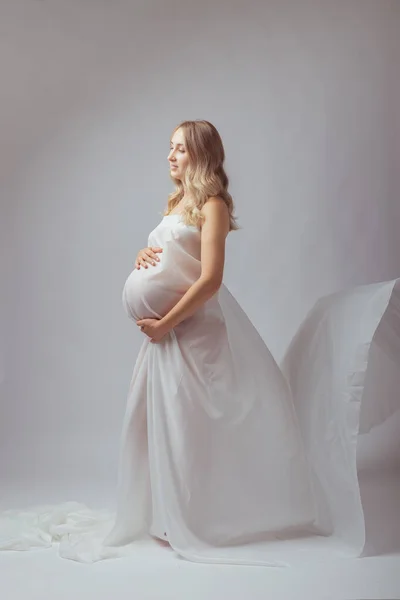穿着飘扬的白色面料站在那里的优雅的孕妇。怀孕、生育和母性概念. — 图库照片