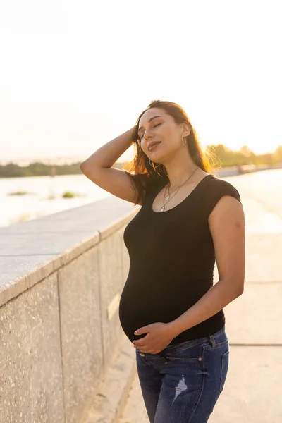 Porträtt av en lycklig gravid latinamerikansk kvinna som tittar på sidan promenader i en park vid solnedgången. Graviditet och moderskap koncept. — Stockfoto