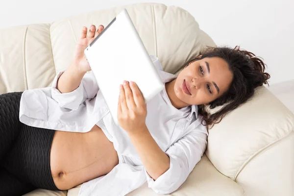 Latim mulher grávida usando computador tablet enquanto deitado no sofá em casa. Gravidez e informação para o conceito de paternidade. — Fotografia de Stock