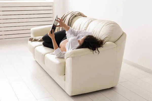 Латинська вагітна жінка, лежачи на дивані, користується планшетним комп "ютером. Вагітність та інформація про батьківство. — стокове фото