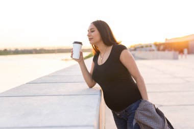 Young pregnant woman drinking takeaway coffee or tea on embankment. Maternity, leisure and health concept. Copy space.