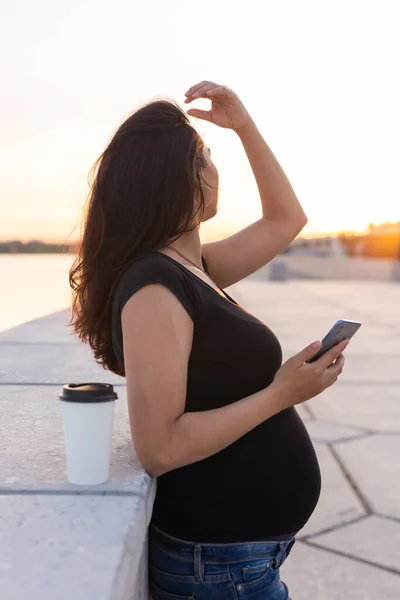 Gravid latinamerikansk kvinna håller smartphone och koppla av på vall med varmt ljus på morgonen — Stockfoto