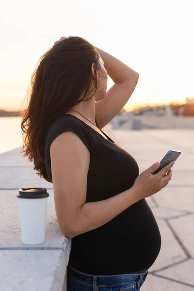Беременная латиноамериканка держит смартфон и расслабляется на набережной с теплым светом утром — стоковое фото