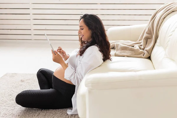 Latin gravid kvinna med hjälp av surfplatta dator sitter nära soffan hemma. Graviditet och information för föräldraskap koncept. — Stockfoto