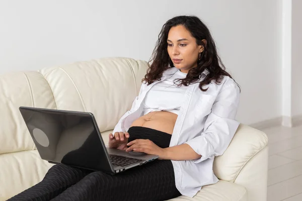 Латиноамериканская беременная женщина с ноутбуком сидит дома на диване. Беременность и информация о концепции родительства. — стоковое фото